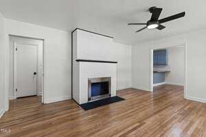 A bright, open room with polished hardwood floors and white walls. A white brick fireplace with a black mantle stands in the corner. A black ceiling fan is in the center of the room, and two doorways lead to adjacent spaces.