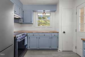 A kitchen with light blue cabinets, a wooden countertop, and a black oven with a towel hanging on the handle. There's a window above the counter letting in natural light, alongside a white door and gray flooring.