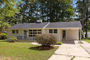 Nestled at 215 Pine Tree Road, this charming Oxford home is a white single-story retreat with a dark roof. Encased by trees, it boasts large windows, a welcoming front porch with railing and an attached carport. A driveway leads to the house, framed by shrubs and a lush green lawn.
