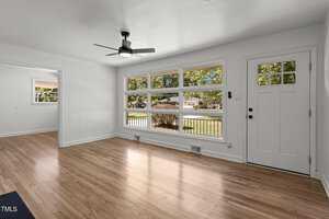 Bright living room with large windows, light wood flooring, and a ceiling fan. The space opens to a room on the left, while a white front door with glass panels is on the right. Outside, greenery is visible through the windows.