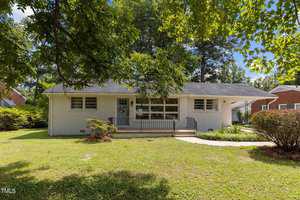 A single-story white brick house with a dark roof, surrounded by trees and greenery. The house has a small front porch with a wreath on the door, and a curved walkway leading to the entrance. The yard is grassy and bordered by shrubs.