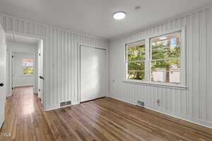 A small, bright room with white vertical panel walls, wooden floor, and a window showing green trees outside. A closed white door leads to another area. The space has a clean, simple design.