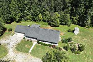 Aerial view of a single-story house with a gray roof surrounded by lush greenery and trees. A small shed is visible in the backyard, and a pathway leads to the front door. The yard is spacious, with a mix of grass and bushes.