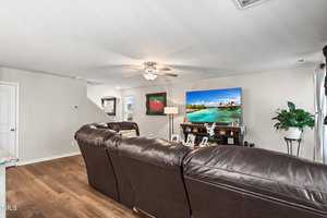 A cozy living room with brown leather couches facing a large TV displaying a tropical beach scene. A ceiling fan is above, and a staircase is visible on the left. There's a potted plant in the corner, and family photos are on a wooden cabinet under the TV.