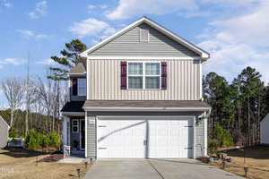 A two-story house with light gray siding, white garage door, and maroon shutters. The driveway leads to a two-car garage. The yard is neatly manicured, and trees are visible in the background, under a blue sky with a few clouds.