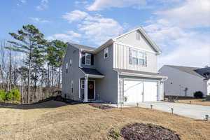 Two-story gray house with a two-car garage, set on a sloped lawn. A wreath decorates the front door. Tall trees are visible in the background under a partly cloudy sky.