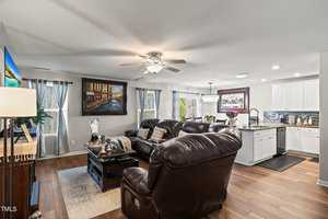 A cozy living room with brown leather sofas facing a wall-mounted TV. There's a coffee table with decor, hardwood floors, and large windows with curtains. The open kitchen has white cabinets, stainless appliances, and a colorful backsplash.