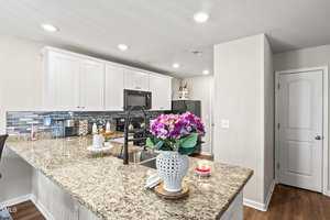 Modern kitchen with granite countertops, white cabinets, and stainless steel appliances. A bouquet of purple and pink flowers is on the island, along with a candle. The backsplash features a gray tile design. Wood flooring complements the space.