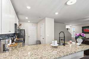 Modern kitchen with granite countertops, stainless steel appliances, and white cabinetry. A black faucet stands out on the island. In the background, there's an open living area with a brown leather couch and a vase of purple flowers.