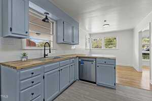 A modern kitchen with light blue cabinets, wood countertops, and stainless steel appliances. The room features a large window with natural light, a potted plant by the sink, and a wooden floor extending into an adjoining room.