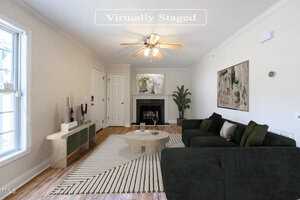 A virtually staged living room featuring a green sectional sofa, round wooden table, abstract rug, and a fireplace. A tall plant and modern decor items are placed along the walls. A ceiling fan is centered above. Natural light enters through the window.