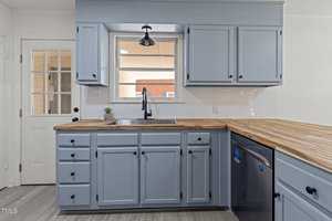 A kitchen with light blue cabinets, a wooden countertop, and stainless steel sink. The backsplash is white tile, and there's a window over the sink. A door with a window and a dishwasher are visible on the left and right, respectively.