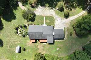 Aerial view of a house with a dark roof, surrounded by green grass and trees. The property includes a small outbuilding and a gravel driveway curving around the front. A pathway leads to the front entrance.