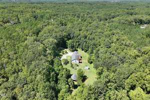 Aerial view of a secluded house surrounded by dense green forest. The property includes a main house, a smaller structure, and a few open spaces in a clearing, with a driveway leading through the foliage.