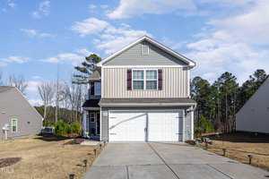 The two-story house at 1570 Dorsett Lane in Creedmoor features gray siding and a white garage door, nestled in a suburban neighborhood. A well-maintained lawn complements the concrete driveway, while pine trees and a partly cloudy sky create a serene backdrop.