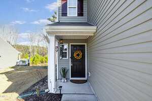 Front entrance of a gray house with a covered porch, featuring a black door adorned with a sunflower wreath. The walkway is lined with small lights, and the yard is visible with a clear blue sky in the background.