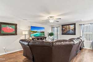 A cozy living room features a large curved leather sofa facing a flat-screen TV displaying a tropical beach scene. Paintings adorn the walls, and a ceiling fan provides overhead lighting. Natural light streams in through two windows with curtains.