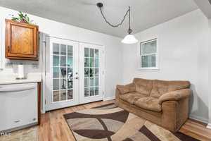 A cozy room with a brown sofa on a patterned rug, set near a pair of French doors leading outside. The walls are white, and there's a hanging light fixture. A small window and a wooden cabinet are visible in the corner.
