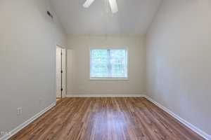 An empty room with light-colored walls, a vaulted ceiling, and a ceiling fan. The hardwood floor leads to a window with blinds, letting in natural light. A doorway to the left reveals a glimpse of another room.