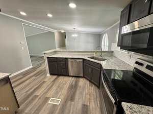 Modern kitchen with dark cabinets, granite countertops, and stainless steel appliances. The open layout leads to a well-lit living area with a chandelier. Light wood flooring and a window add to the ambiance.