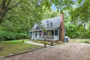 A charming blue house at 106 Stratford Court, with a wraparound porch, nestled among lush trees. It features a brick chimney and an American flag. The front yard is well-maintained with small shrubs, and a gravel driveway in Oxford leads to the entrance.