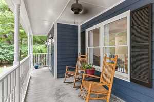 A wooden porch with two rocking chairs and a potted plant. The porch is attached to a blue house with white trim and black shutters. A ceiling fan is installed above. Trees and greenery are visible in the background.