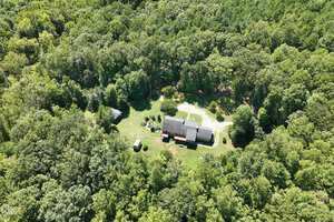 Aerial view of a house at 4161 Fernwood Trail, nestled in lush green forest near Oxford. The main building and two smaller structures are partially shaded by trees, with a grassy clearing and dirt path visible in the foreground.