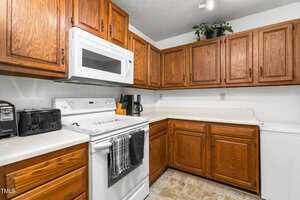 A kitchen with wooden cabinets, white countertops, an electric stove, and a microwave. A coffee maker and a toaster are on the counter. A plant decorates the upper cabinet. The floor has a tiled pattern.