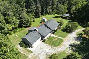Aerial view of a single-story house with a dark roof, surrounded by lush greenery and trees. There is a gravel driveway leading to the garage. A small structure and a gazebo are in the backyard.