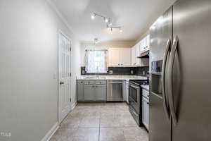 A modern kitchen with stainless steel appliances, including a fridge and oven. The cabinets are white and gray, and there's a window with sheer curtains. The floor is tiled, and the lighting is bright with ceiling spotlights.