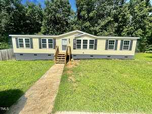 A yellow single-story modular home at 6611 Clearwater Drive, with a gray foundation, sits on a grassy yard in Oxford. A wooden staircase leads to the front door, framed by several large windows. Trees sway in the background under a clear blue sky.