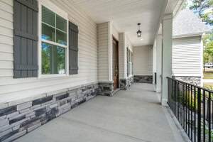 A modern porch with stone accents on the walls, white siding, and dark shutters. It features a wooden front door, black railing, and overhead lighting. The porch floor is concrete, and there are trees visible in the background.