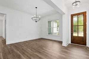A bright, empty room with white walls, wooden flooring, and a wooden door. A large window with blinds is on the right wall. A modern black chandelier hangs from the ceiling. An open doorway leads to another room on the left.