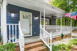 A cozy house features a blue exterior with white trim, a covered front porch with railing, and two lantern-style wall lights. An American flag is mounted on the porch, and there are plants in the garden beside the brick steps.