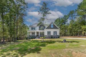 A two-story house with light siding and dark roof is surrounded by tall trees. It features a front porch with columns and a grassy yard under a partly cloudy sky. A stone is near the house on the right side of the lawn.
