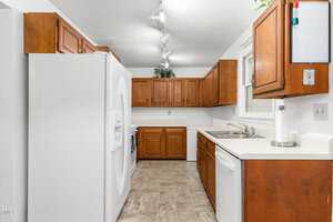 A kitchen with wooden cabinets, white countertops, and tile flooring. It features a white refrigerator, dishwasher, and a sink under a window. Overhead lighting and a small potted plant on the cabinet add to the decor.