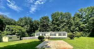 A gray manufactured home with white trim sits on a spacious green lawn, surrounded by tall trees under a clear blue sky. A pathway leads to the front steps, and a small yellow shed is visible to the left.