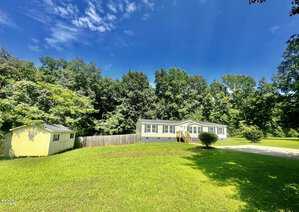 A small, single-story house with a light exterior is set against a backdrop of lush trees under a blue sky. A wooden deck leads to the entrance. A yellow shed sits on the left, and a large grassy lawn surrounds the structures.