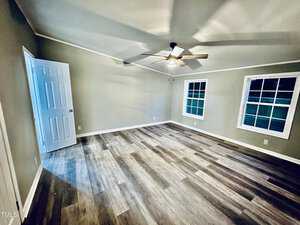 An empty room with light wood laminate flooring and beige walls. It features a ceiling fan with lights, two windows with white trim, and an open white door leading to another space.
