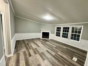 A spacious living room with light gray walls and wood-paneled flooring. There is a white fireplace against the far wall, beneath a ceiling light. Three windows are on the right, letting in natural light. A white chair rail runs around the room.