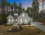 A modern two-story house at 1119 Springdale Drive in Wake Forest boasts a light exterior, surrounded by a manicured lawn and trees. The sunset sky's pink and blue hues add warmth to the serene landscape, while a driveway leads to the side garage.