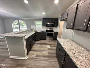 A modern kitchen with dark cabinets, granite countertops, and stainless steel appliances. The space is well-lit with recessed lighting and features a large window letting in natural light. The floor has a wood-like finish.