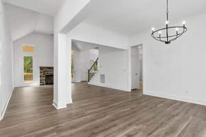 Spacious empty room with light hardwood floors, tall white walls, and large windows. A modern black chandelier hangs from the ceiling. A stone fireplace is visible in the background, along with a staircase. Natural light fills the space.