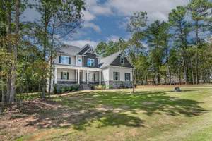 A large two-story house with white siding and stone accents is surrounded by a grassy yard and tall trees. The sky is partly cloudy, creating a serene and picturesque outdoor setting.