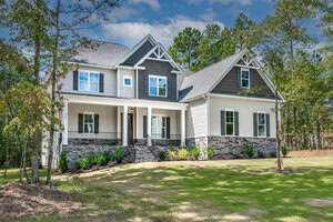 A two-story house with gray and white siding and a stone facade. It has a covered porch, pointed gables, and a well-maintained lawn with surrounding trees under a partly cloudy sky.