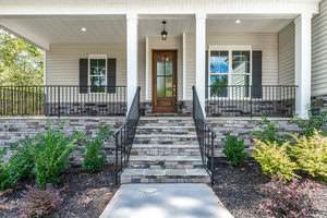 A front view of a house entrance with stone steps leading to a wooden door. The porch is framed with white columns, and two windows with shutters are on either side. The exterior has stone and siding details, surrounded by neatly trimmed bushes.
