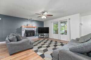 A cozy living room with a geometric-patterned rug, two gray couches, and a large flat-screen TV. A brick fireplace decorates the blue accent wall. Natural light streams through a window overlooking a green outdoor space. Ceiling fan and wooden floor.