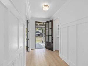 A bright, minimalist entryway with white walls and light wood flooring. A large glass door is open, revealing a grassy lawn and a suburban house in the background. A modern ceiling light fixture is centered above.