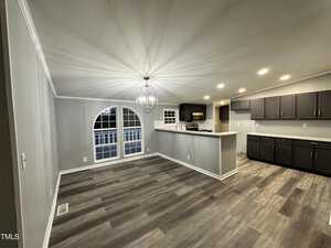 A modern kitchen and dining area with dark cabinets and stainless steel appliances. The room features gray walls, wood-style flooring, and a large decorative light fixture. An arched window provides natural light from the left side.