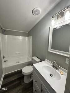 A small bathroom featuring a bathtub with a shower, a toilet, and a white vanity sink with a mirror above. The walls are painted gray, and the floor has dark wood-like flooring. A ceiling vent and a light fixture with two bulbs are visible.